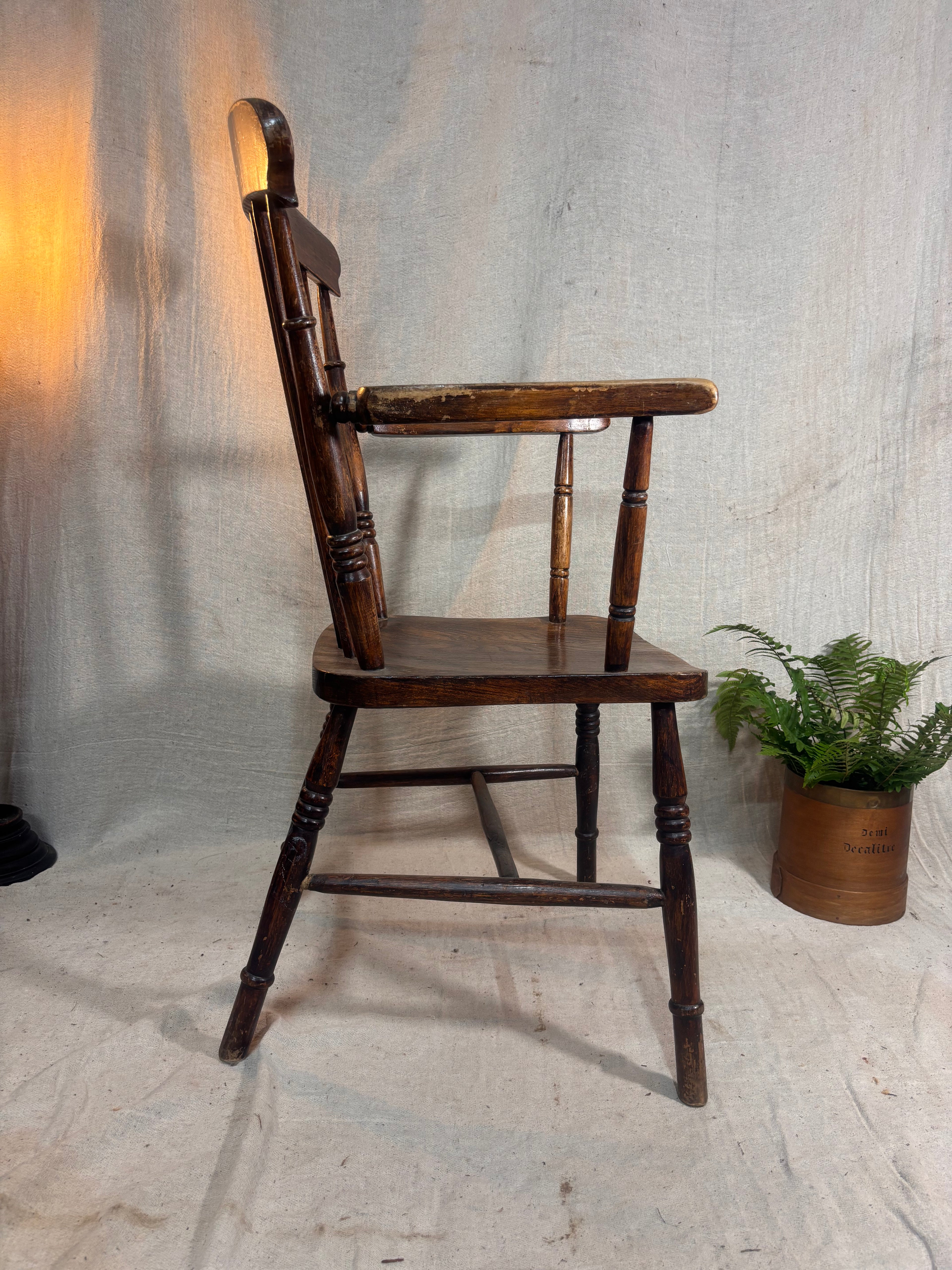 Wooden chair with armrests against a textured wall with a plant in the corner.