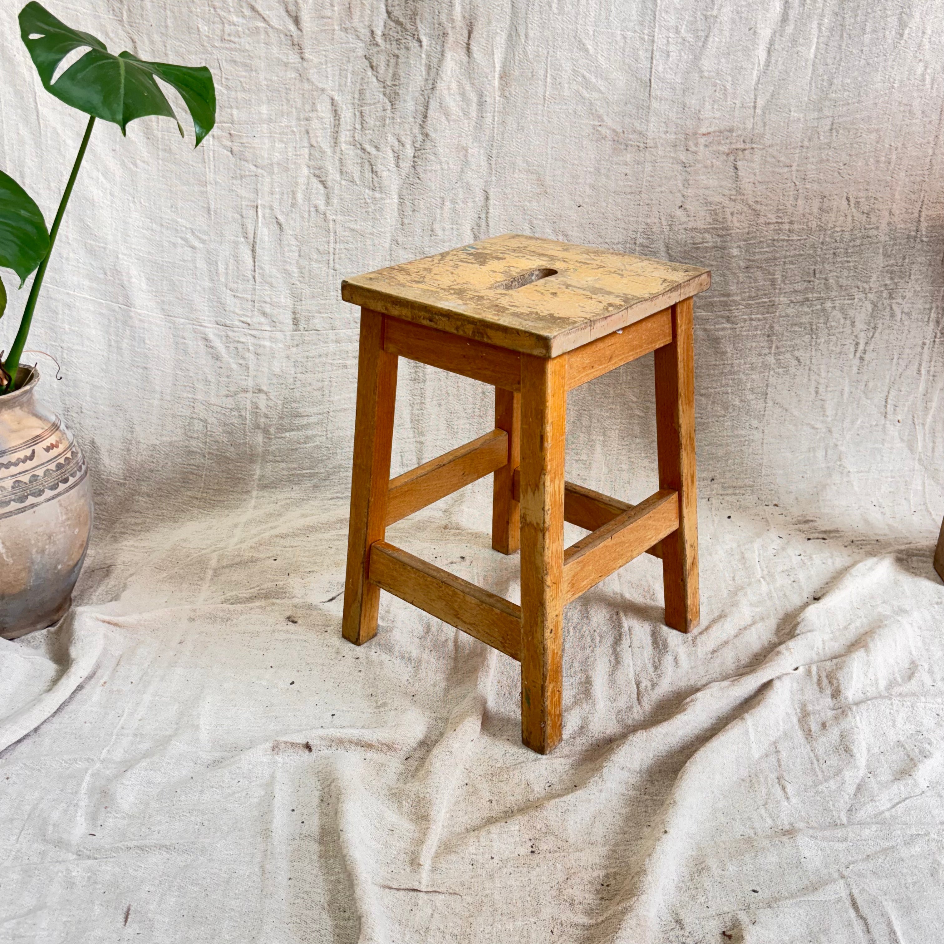 Wooden stool on a textured white surface with a plant in the corner.